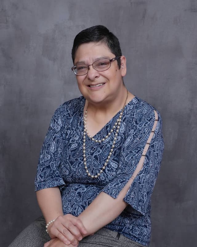 Rebeca Aguirre, creator of the UXAF methodology, seated and smiling warmly in a professional portrait. She wears glasses, a navy paisley top, layered pearl necklaces, and a pearl bracelet. A hearing aid is visible behind her ear. Her expression is confident and approachable.